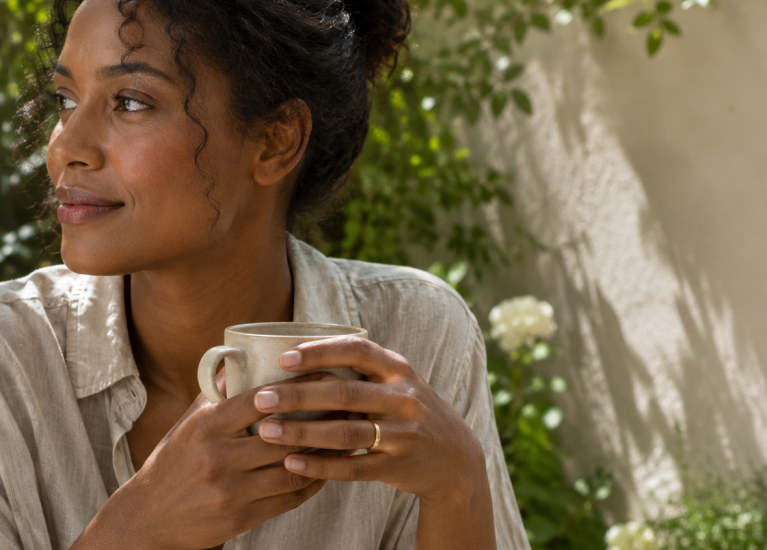 Woman wearing a Phoebe 2mm yellow gold wedding band, holding coffee at a garden table